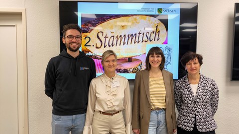 Gruppenbild mit 4 Personen vor einem großen Bildschirm