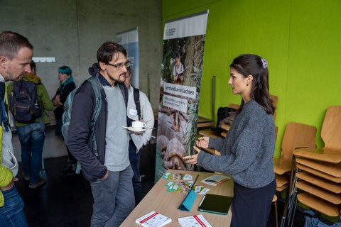 Mann und Frau unterhalten sich während im Hintergrund buntes Treiben herrscht.