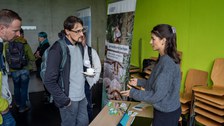 Mann und Frau unterhalten sich während im Hintergrund buntes Treiben herrscht.