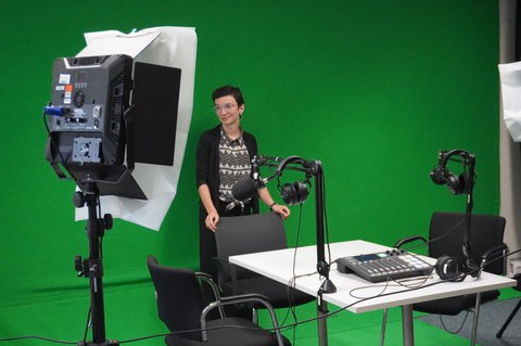 Frau steht vor Greenscreen und vor ihr ist Podcast-Technik zu sehen