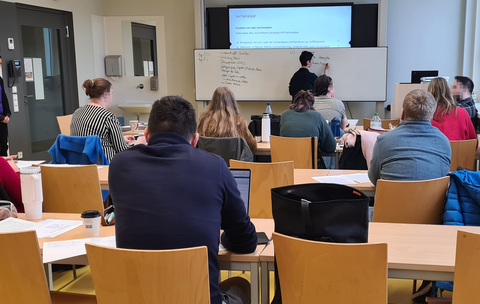 Frau schreibt während Vortrag etwas ans Whiteboard im Seminarraum. Seminarteilnehmende von hinten fotografiert.