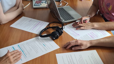 Nahaufnahme eines Tisches mit Notizen, Laptop und Kopfhörern. 4 Personen sitzen an diesem Tisch und arbeiten gemeinsam