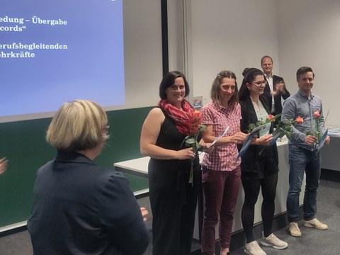 Vier Personen stehen vor einer Tafel mit jeweils ihren Transcript of Records und einer Rose in der Hand.