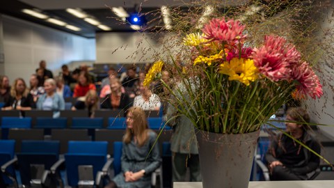 Blumenstrauß auf Tisch, im Hintergrund sitzen Personen im Hörsaal