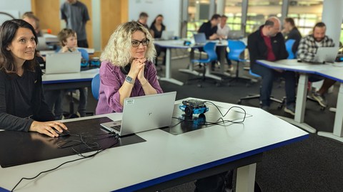 Gruppe von Erwachsenen sitzen in einem Seminarraum und haben Laptop und kleinen Roboter neben sich