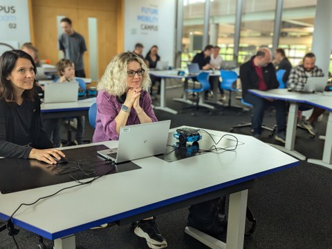 Gruppe von Erwachsenen sitzen in einem Seminarraum und haben Laptop und kleinen Roboter neben sich