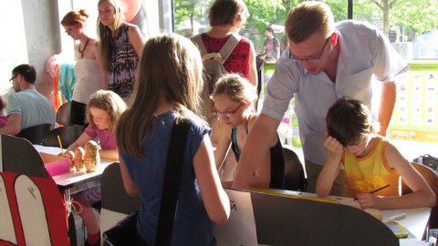 Bild mit mehreren Personen, darunter ein Großteil Kinder, die teilweise an einem Tisch sitzen, teilweise daneben stehen und malen, zeichnen oder zuschauen.