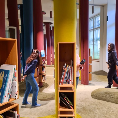 Ein kleines Bücherregal hängend an einer Säule ist im Vordergrund zu sehen. Im Hintergrund ist eine Lernwerkstatt zum Thema Bilderbücher zu sehen - bunte Säulen, Teppichboden, weitere Bücherregale