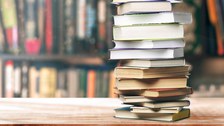 Bild mit einem Stapel verschachtelt aufeinander liegender Bücher auf einer Holzplatte. Im Hintergrund ist verschwommen ein Regal mit weiteren Büchern zu sehen.