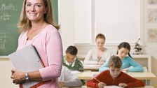 Bild, auf dem eine Lehrerin mit einem Notizbuch in einem Klassenzimmer steht. Im Hintergrund ist ein Teil einer Kreidetafel und Bankreihen mit jungen Schüler:Innen zu sehen.