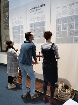 Bild von drei Personen, die vor einer Informationsgrafik über Seemannsknoten stehen. Sie lesen diese und versuchen gleichzeitig mit ihren Händen die vor ihnen hängenden Seile zu verknoten.