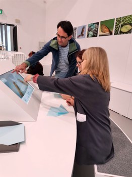 Bild, auf dem zwei Frauen und ein Mann vor einem Touchscreen sitzen bzw. stehen und auf den Bildschirm zeigen.