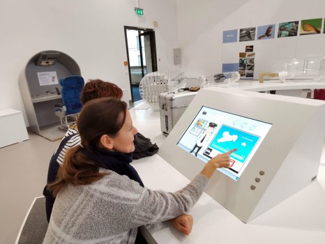 Bild, auf dem zwei Personen in einem überwiegend weißen Raum vor einem Touchscreen sitzen, wobei eine der beiden Personen eine Auswahl mit ihrem Finger trifft.