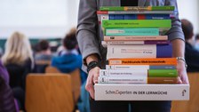 Bild, auf dem eine Person mit einem Stapel Büchern in den Händen in einem Hörsaal steht. Im Hintergrund sind verschwommen eine Tafel und Stühle zu sehen, auf denen mehrere Personen sitzen.