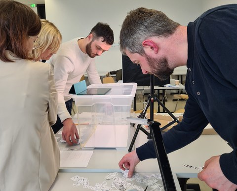 Personen betrachten im linken Hintergrund die Aufnahmemöglichkeiten auf einem Tablet, welches sich auf einer transparenten Kiste befindet. Eine weitere Person im Vordergrund sucht sich Papierfiguren aus.