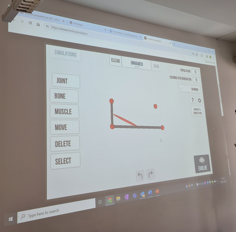 Auf dem Bild sieht man das Beamer-Bild in einem Hörsaal, auf dem Bild wird das Spiel "Evolution" dargestellt, bei dem eine Figur mit der richtigen Kombination an Muskeln und Gliedern zu laufen beginnt.