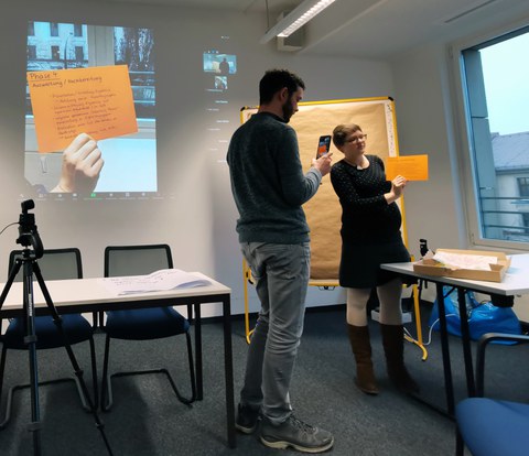 In einem hybriden Galerierundgang präsentiert eine Teilnehmerin (ganz rechts im Bild) zum Abschluss die eigenständig erstellte Lernkiste. Herr Krone (in der Mitte vom Bild) filmt diese Live-Demonstration mit einem Smartphone ab und streamt sie in den virtuellen Raum. Links oben im Bild sieht man das Video, das das Smartphone aufnimmt, auf einer Beamerprojektion.