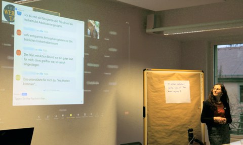 In der rechten Bildhälfte steht die Dozentin Melanie Wohlfahrt, die auf eine Beamer-Projektionsfläche im linken Bildbereich blickt. Hier kann man das Feedback mehrerer Teilnehmender lesen, die sich im Zoom-Chat zur Werkstattwoche wohlwollend äußern.