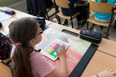 Mädchen sitzt an ihrem Platz im Klassenraum. Auf ihrem Tisch liegt ein Tablet.