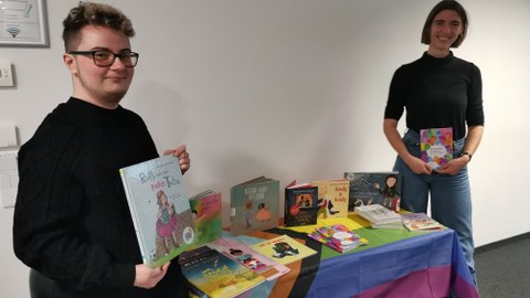 Zwei Personen stehen an einem Tisch gefüllt mit Schulbüchern für queere Grundschulkinder