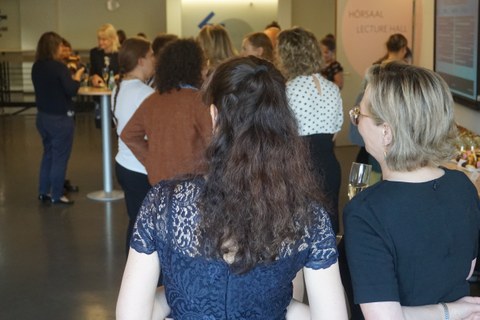 Das Bild zeigt zwei Frauen von hinten, wie sich unterhalten. Im Hintergrund ein Raum zu sehen, wo Personen an einer Abschlussfeier teilnehmen.