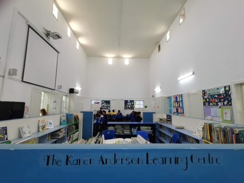 Das Foto zeigt einen großen Raum mit einer blauen Theke und vielen Büchern darin verstaut. Im Hintergrund sind Personen zu sehen, die gerade lernen.