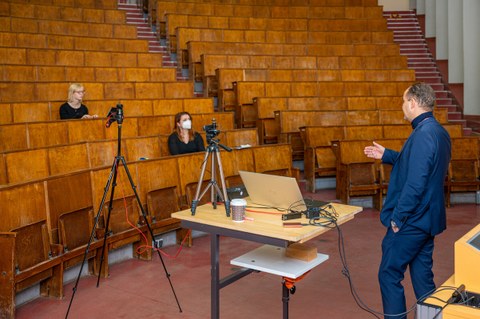 Foto zeigt einen fast leeren Hörsaal mit nur zwei Studentinnen und einen Dozenten, die die digitale Vorlesung aufnehmen.