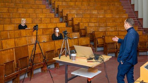 Foto zeigt einen fast leeren Hörsaal mit nur zwei Studentinnen und einen Dozenten, die die digitale Vorlesung aufnehmen.