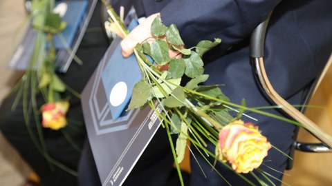 Das Foto zeigt die Hände einer Person im Anzug. Sie hält eine Rose, einen Umschlag und ein Heft in der Hand.