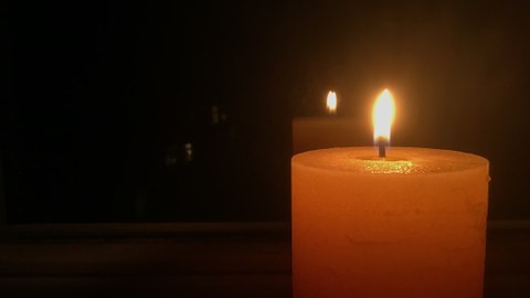 Das Foto zeigt eine breite, leuchtende Kerze in der rechten Bildhälfte in einem sonst schwarzen Bild, die sich im Hintergrund ein einer Reflektion spiegelt.