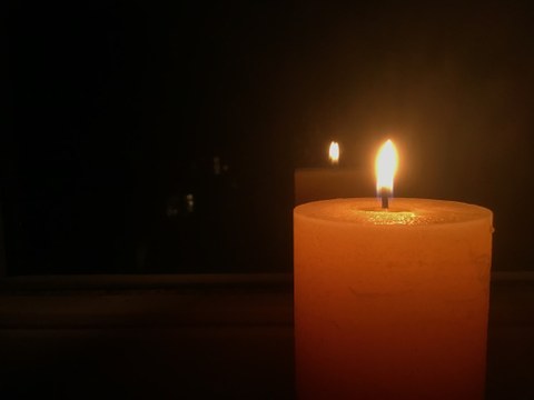 Das Foto zeigt eine breite, leuchtende Kerze in der rechten Bildhälfte in einem sonst schwarzen Bild, die sich im Hintergrund ein einer Reflektion spiegelt.